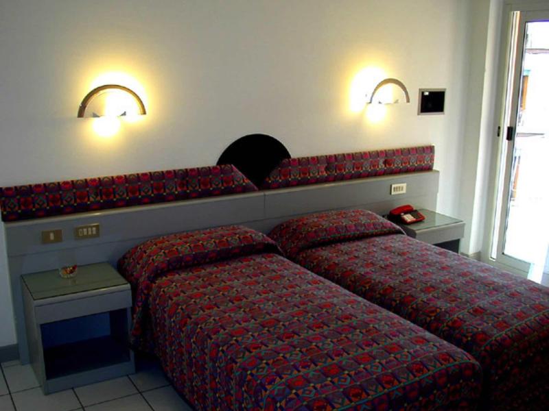 Zwei Einzelbetten mit bunten Decken, Nachttisch, Wandlampen und Balkon in hellem Zimmer.