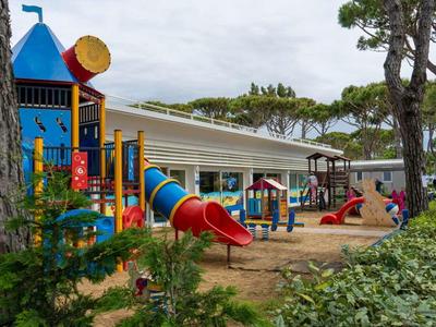 Bunter Kinderspielplatz mit Rutschen, Klettergerüsten und Sandfläche vor einem Gebäude mit Bäumen.