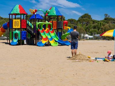 Bunter Spielplatz mit Rutschen am Strand, umgeben von Sand und einigen Schirmen.