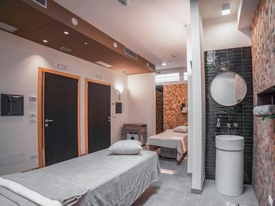 Moderne Behandlungsräume mit weißen und grauen Wänden, zwei Liegen und angrenzendem Waschbeckenbereich.