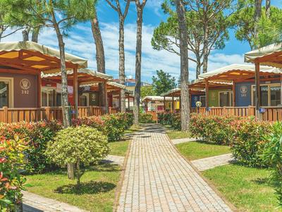 Wanderweg zwischen Holzhütten in grüner Gartenanlage mit Bäumen unter blauem Himmel.