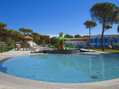Runder Pool mit Wasserrutsche, Sonnenschirmen und blauen Gebäuden im Hintergrund bei klarem Himmel.