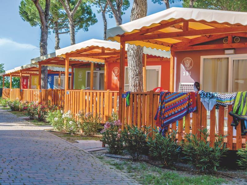 Reihenhäuser mit Holzbalkonen, umgeben von Bäumen, bei klarem Himmel in einer Ferienanlage.