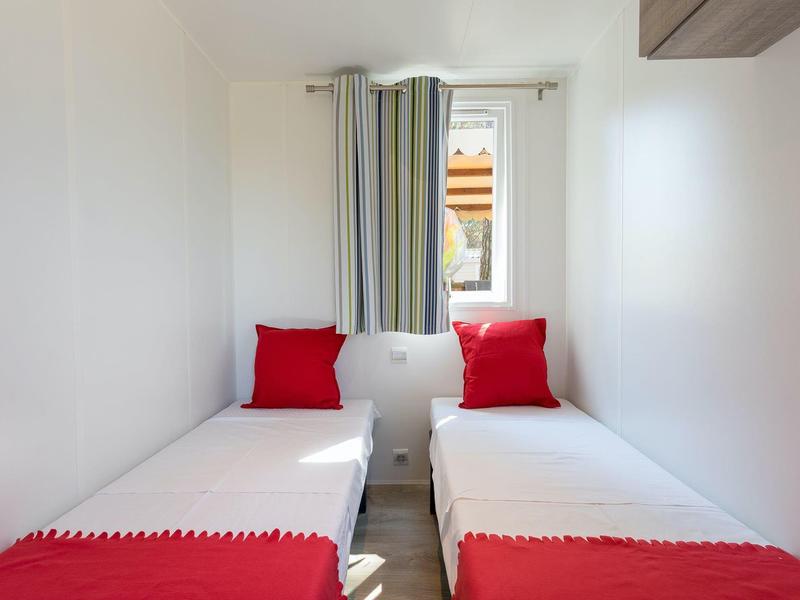 Schmaler Raum mit zwei Einzelbetten, weißen Wänden, roten Kissen und Decken, kleinem Fenster mit Vorhängen.