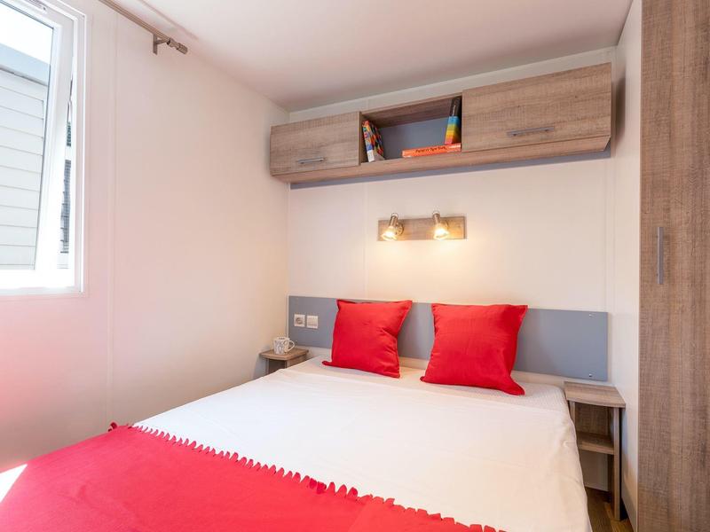 Kleines Schlafzimmer mit Doppelbett, roten Kissen, Holzmöbeln und Fenster rechts.