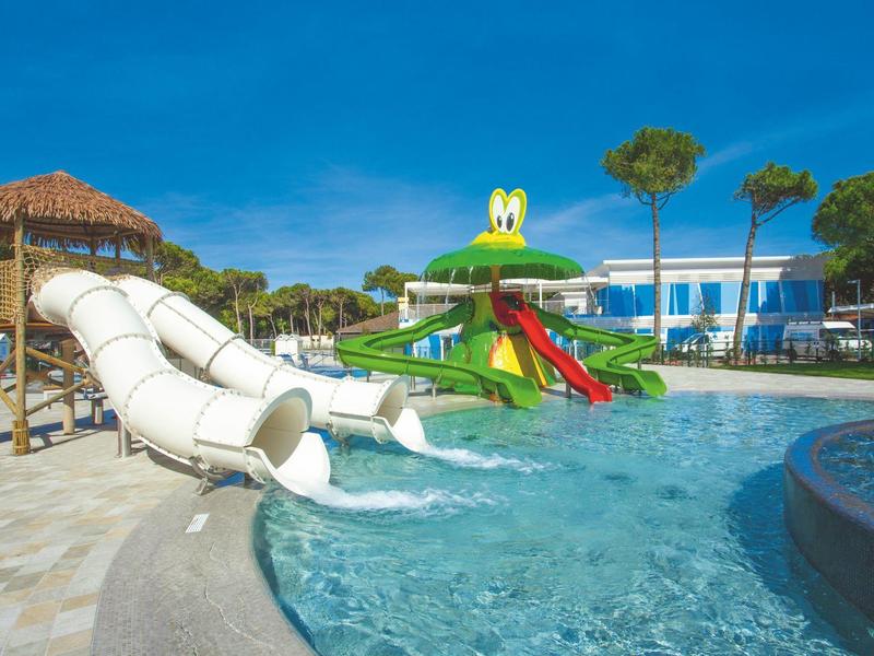 Wasserpark mit zwei Rutschen, einem Planschbecken und einem Spielhaus bei klarem Himmel.