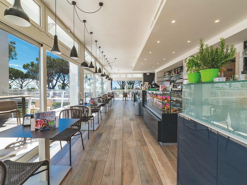 Heller, moderner Café-Innenraum mit Holzboden, schwarzen Stühlen und großen Fenstern zur Terrasse.