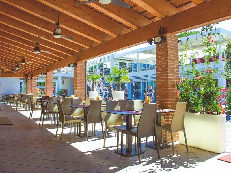 Überdachte Terrasse mit Tischen und Stühlen neben einem Hotelgebäude, sonnig und begrünt.