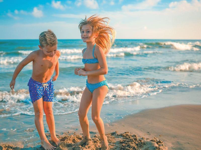 Zwei Kinder spielen am Strand im Sand bei Sonne und leicht bewölktem Himmel.