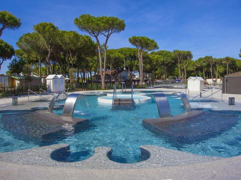 Schwimmbad mit blautürkissem Wasser, drei Metallwasserspendern und dahinter grünen Pinien.