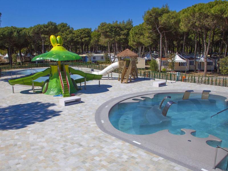 Kinderspielplatz mit grünem Pilz-Wasserspiel und kleinem runden Planschbecken, umgeben von Bäumen.