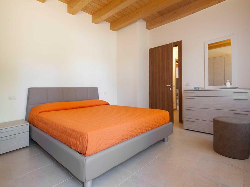 Schlafzimmer mit grauem Bett, orangenem Bettbezug, Kommode und Holzbalkendecke.