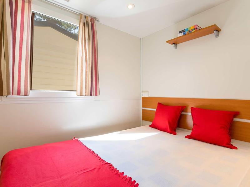 Kleines Schlafzimmer mit weißer Wand, Fenster mit gestreiften Vorhängen, Doppelbett mit rotem Bezug und Kissen.