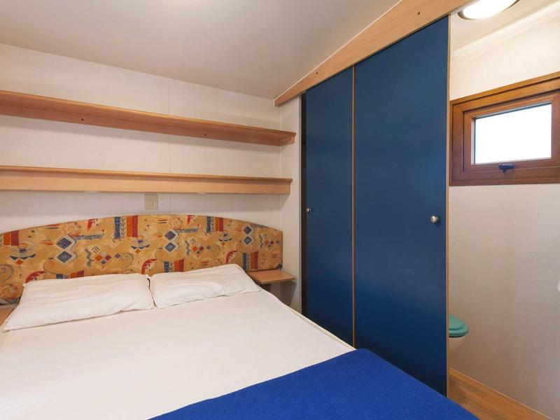 Kleines Schlafzimmer mit Doppelbett, buntem Kopfteil, blauer Schiebetür und hellbraunen Regalen.