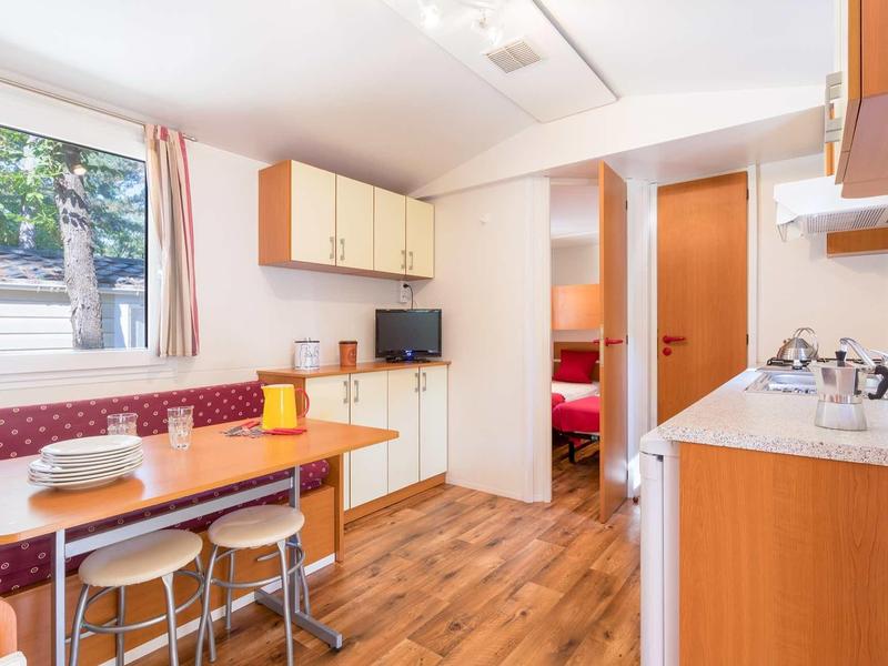 Kleine Wohnküche mit Holzboden, rotem Polstersofa, Esstisch, Küchenzeile und Fenster mit Blick ins Grüne.