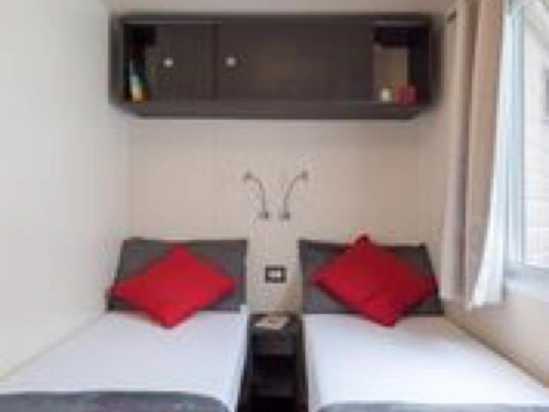 Kleines Zimmer mit zwei Einzelbetten, grauen Bettdecken, roten Kissen und weißem Vorhang am Fenster.