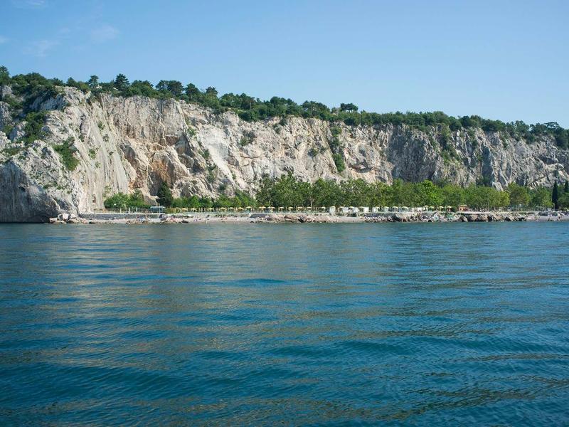 Karge Kalksteinklippe mit grünem Bewuchs und ruhigem blauem Wasser im Vordergrund.