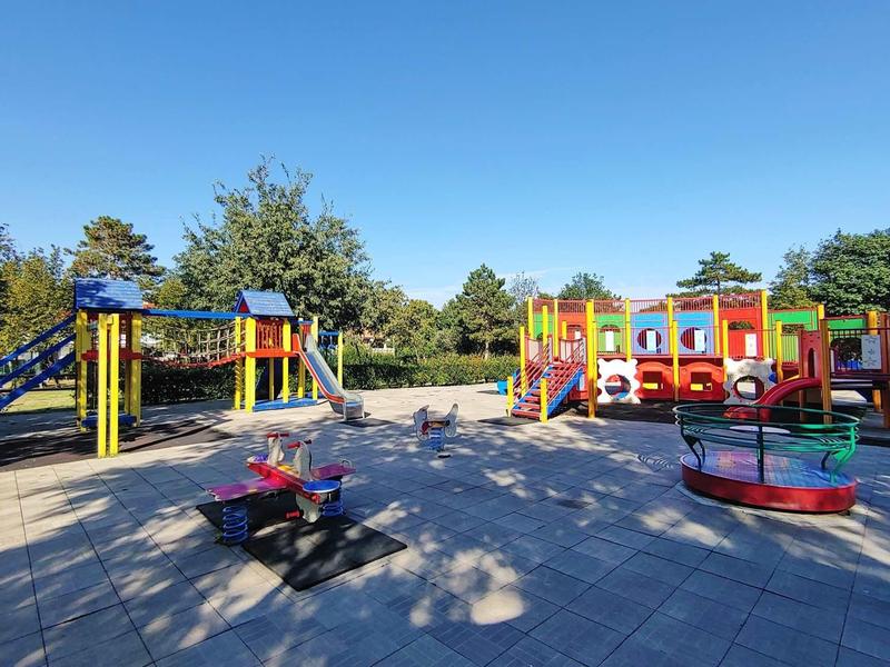 Bunter Spielplatz mit Rutsche, Klettergerüst, Karussell und Sandfläche unter blauem Himmel.