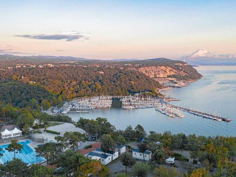 Küste mit Hafen, vielen Booten, grünen Bäumen und einem hellblauen Pool bei Sonnenaufgang.