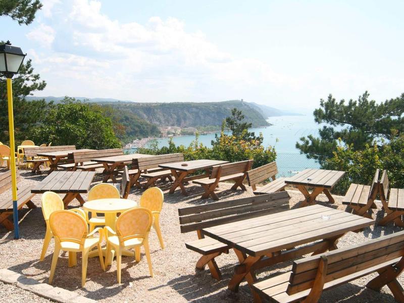 Terrasse mit Holzbänken und Tischen, gelbe Plastikstühle, Blick auf See und bewaldete Berge.