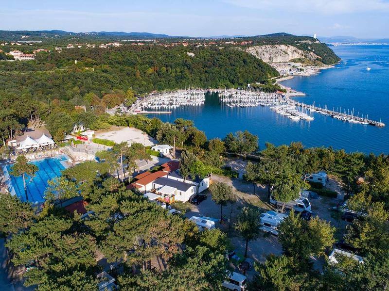 Küstenlandschaft mit grünem Pinienwald, weißen Gebäuden, einem Hafen und blauem Meer unter blauem Himmel.