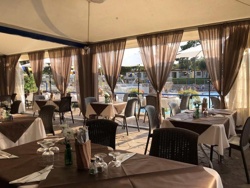 Großes, helles Restaurant mit durchsichtigen Vorhängen und Tischen mit weißen Tischdecken am Wasser.