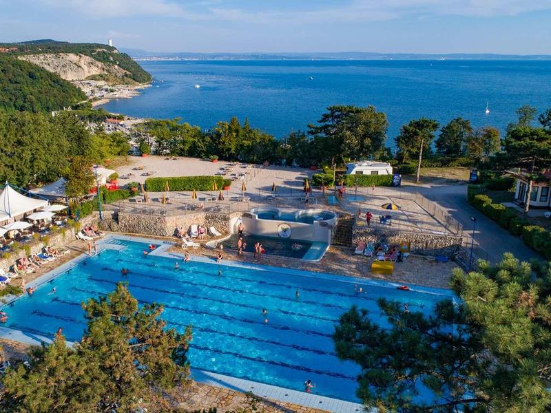 Großes Freiluft-Schwimmbecken in Küstennähe mit umliegenden Bäumen und blauem Meer im Hintergrund.