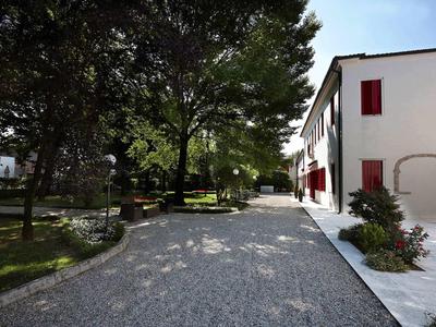 Sonniger Weg mit Bäumen neben einem weißen Gebäude mit roten Fensterläden.