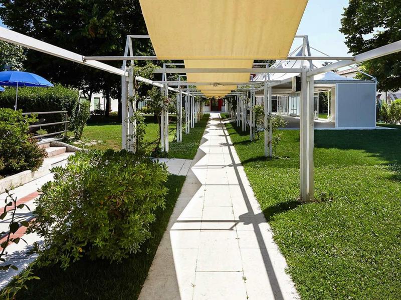 Cieniowany wybrukowany chodnik z żółtym daszkiem i zielonym trawnikiem obok niebieskich parasoli.