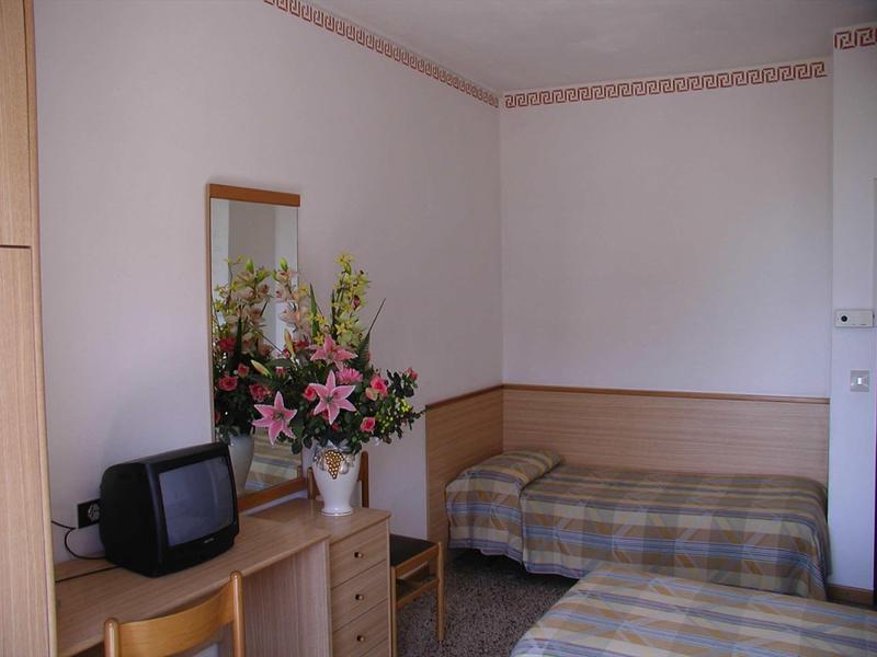 Kleines Zimmer mit Einzelbett, Holzmöbeln, altem Fernseher und Blumenstrauß auf Kommode.
