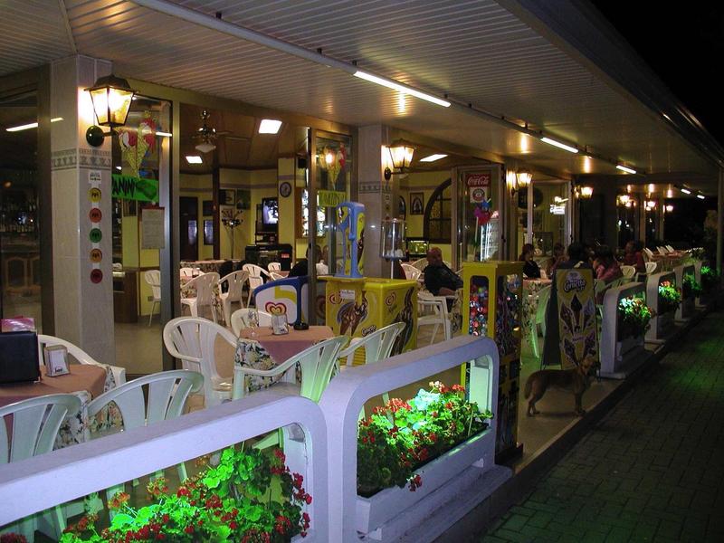 Beleuchtete Außenterrasse eines Restaurants mit weißen Tischen und Stühlen bei Nacht.