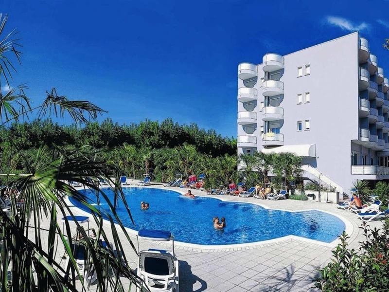 Moderner Hotelkomplex mit rundem Außenpool, umgeben von Palmen und blauem Himmel.