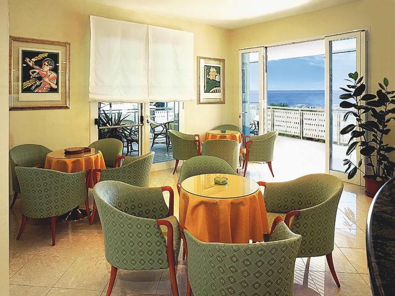 Gemütlicher Sitzbereich mit grünen Sesseln und orangenen Tischtüchern, Meerblick durch große Fenster.