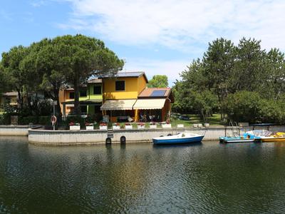 Haus am Wasser mit Booten und Bäumen unter blauem Himmel