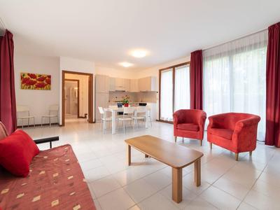 Helles Wohnzimmer mit roten Sesseln, Sofas und einem Holztisch, großen Fenstern mit roten Vorhängen.