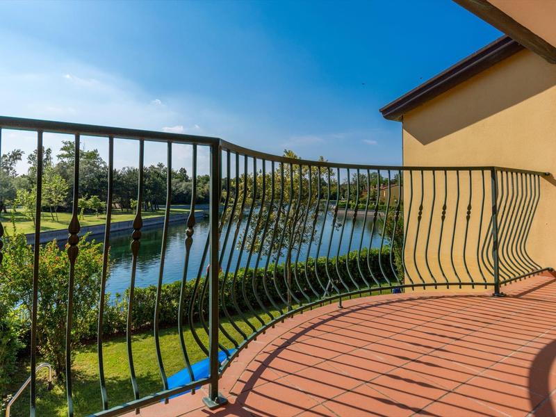 Balkon mit geschwungenem Metallgeländer und Blick auf einen Teich und Bäume bei klarem Himmel.