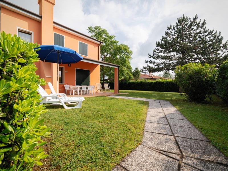 Gartenbereich mit Liegestühlen und Sonnenschirm neben einem zweistöckigen Haus bei bewölktem Himmel.