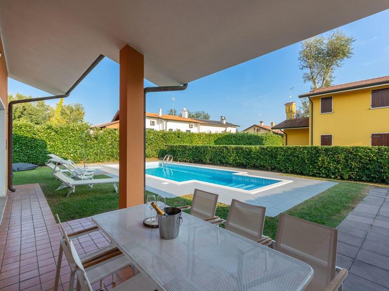 Überdachte Terrasse mit Tisch und Stühlen, Blick auf Pool und Liegestühle im Garten.