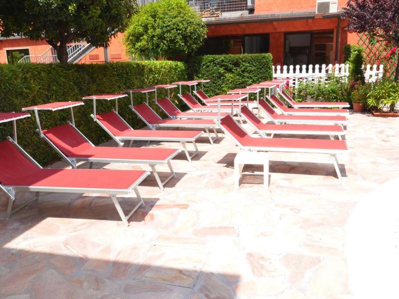 Mehrere rote Liegestühle stehen in einer sonnigen, gepflasterten Außenanlage neben einem Garten.