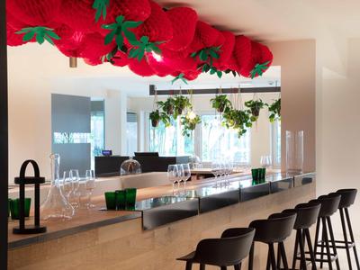 Espace bar d'hôtel moderne avec tabourets, étagères en verre et décorations rouges suspendues.