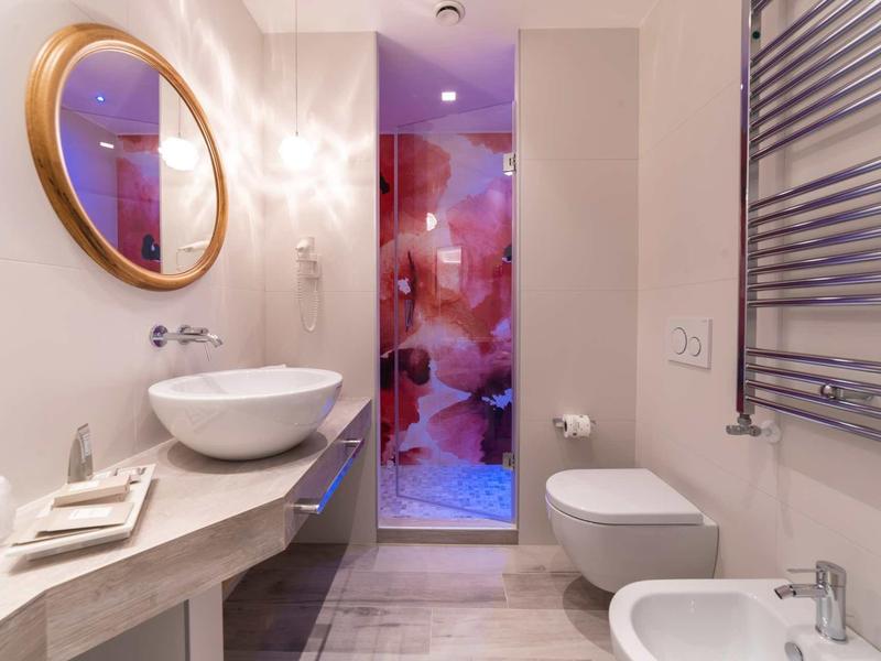 Modernes Badezimmer mit rundem Spiegel, Waschbecken, Toilette und Glasdusche mit farbigem Muster.