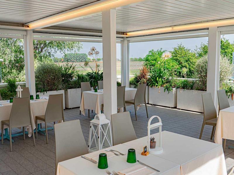 Heller, offener Außenbereich mit weißen Tischen, grauen Stühlen und grüner Pflanzenkulisse.