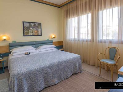 Jasny pokój hotelowy z podwójnym łóżkiem, stolikami nocnymi, krzesłem, telewizorem i dużymi oknami z zasłonami.