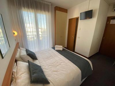 Helles Hotelzimmer mit Doppelbett, weißen und grauen Bettwaren, kleinem Fernseher und Tür zum Balkon.