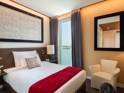 Modernes Hotelzimmer mit Doppelbett, roter Decke, Sessel und großem Fenster mit Vorhängen.