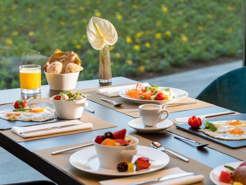 Frühstückstisch im Hotel mit Orangensaft, Kaffee, frischem Obst und Brötchen vor einer großen Fensterfront.