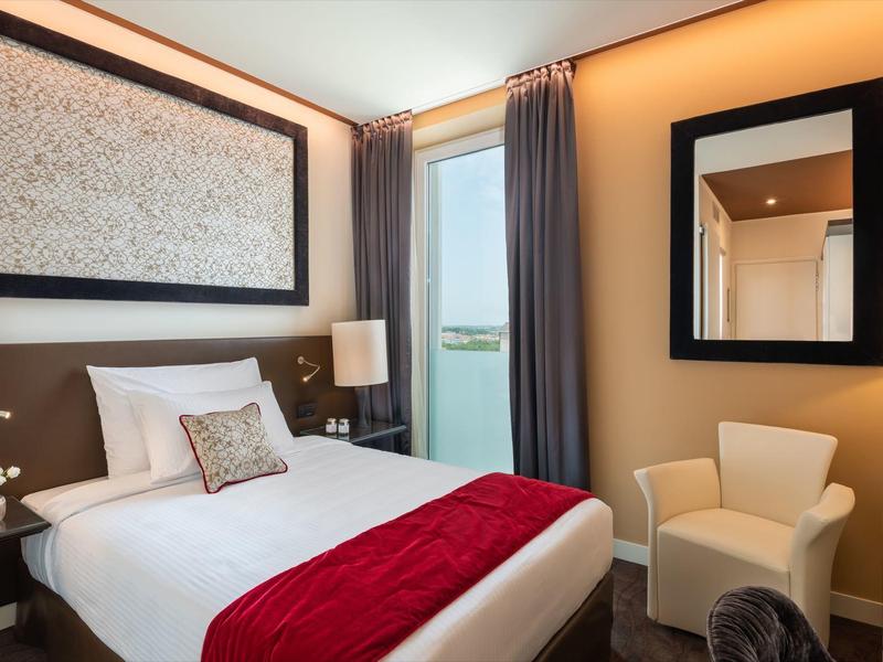 Modernes Hotelzimmer mit Doppelbett, roter Decke, Sessel und großem Fenster mit Vorhängen.