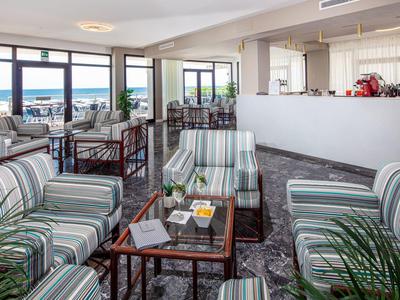 Ampia lounge dell'hotel con poltrone a righe, tavoli e vista mare attraverso grandi finestre.