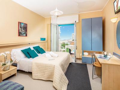 Camera d'hotel luminosa con letto matrimoniale, armadio blu, scrivania e balcone con vista sulla città.