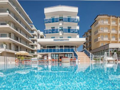 Hotel moderno con piscina e ombrelloni, circondato da altri edifici sotto un cielo sereno.